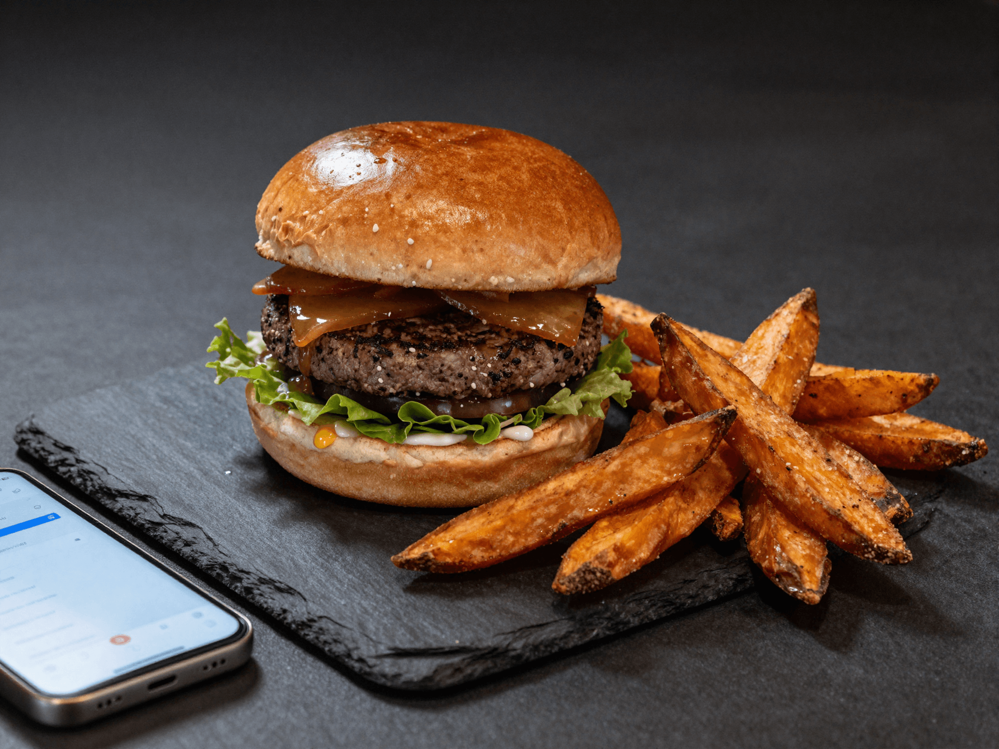 Gourmet burger with sweet potato fries on a dark slate plate, a smartphone next to it